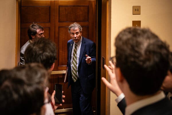 Senator Sherrod Brown stands in an open elevator, talking to reporters standing outside before the door closes.