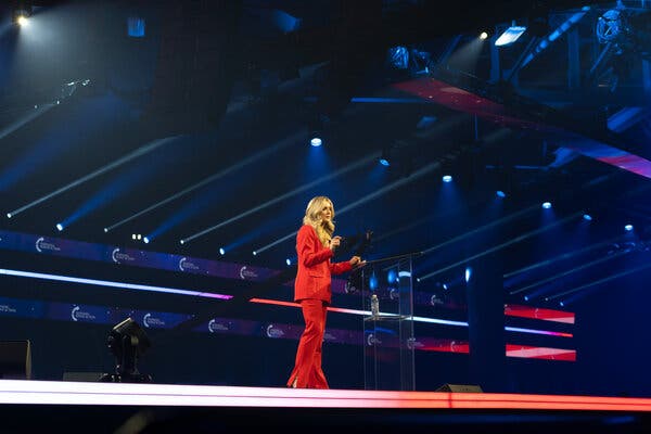 Riley Gaines stands on a stage next to a clear podium, dressed in red.